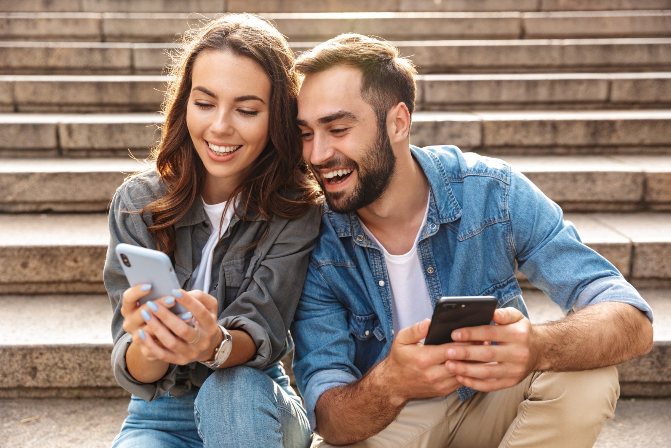 Eine junge Frau und ein junger Mann sitzen auf einer Treppe und buchen im Smartphone eine Freizeitversicherung.